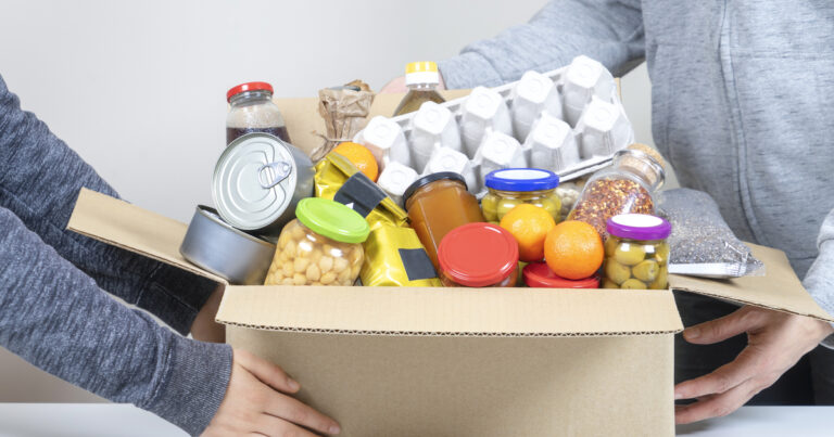 Volunteers hands holding food donations box with grocery products on white desk