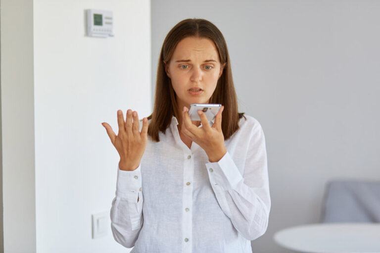 Portrait of confused puzzled female wearing white shirt posing at home with smart phone in hands, raising arm, does not understand why device not working.